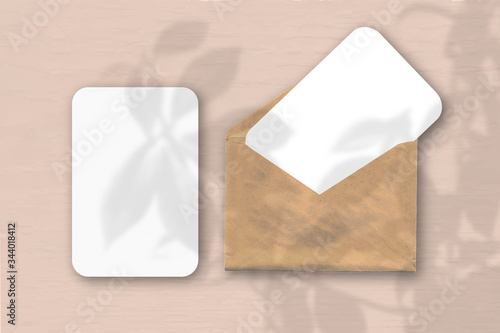 An envelope with two sheets of textured white paper on a pink table background. Mockup with an overlay of plant shadows. Natural light casts shadows from a tropical plant.