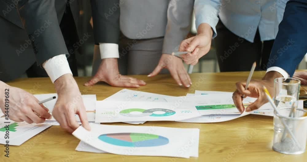 Close up of hands mixed-races people working with papers and documents with charts, graphics and diagrams lying on wooden desk in office. Business meeting and discussion concept Multiethnic colleagues Close up of hands mixed-races people working with papers and documents with charts, graphics and diagrams lying on wooden desk in office. Business meeting and discussion concept Multiethnic colleagues
