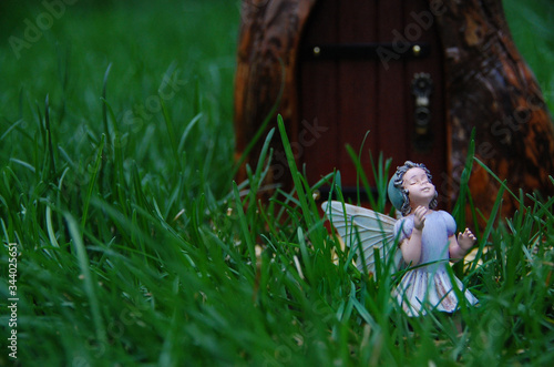 Sweet fairy statue stands in fairy garden surrounded by green grass. Fairy princess is in front of door to magical fantasy world. She is beautiful, mystical, and delicate. © Katherine
