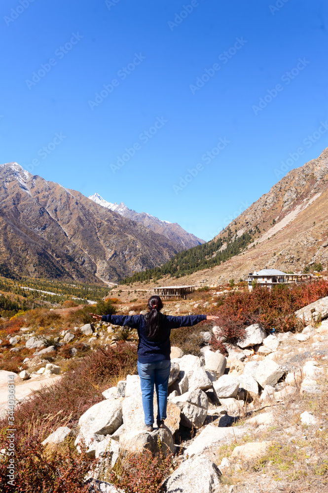 Obraz premium Solo Woman Indian traveler in winter casual standing alone with arms outstretched in mountain valley. Snow capped Himalayan mountain forest blue sky in background. Spiti Valley Himachal Pradesh India 
