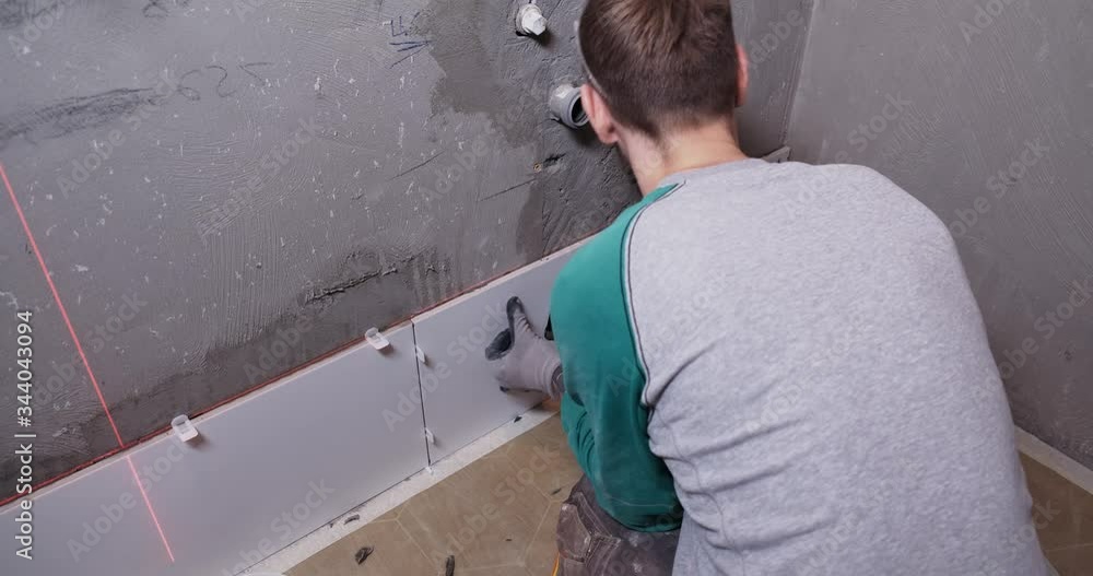 Male tiler put ceramic tiles on wall in bathroom, laser level highlights place of installation, professional approach, adhesive is applied to wall, builder presses tile to wall, exact, repair in house Male tiler put ceramic tiles on wall in bathroom, laser level highlights place of installation, professional approach, adhesive is applied to wall, builder presses tile to wall, exact, repair in house