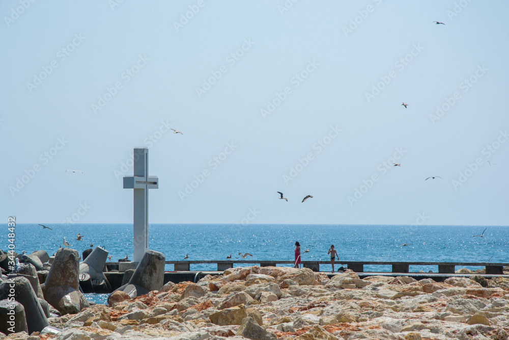  Seascape, view of a big concrete cross on the bay, people walking and birds flying, blue waters and sky, breakwaters, summer season, nature