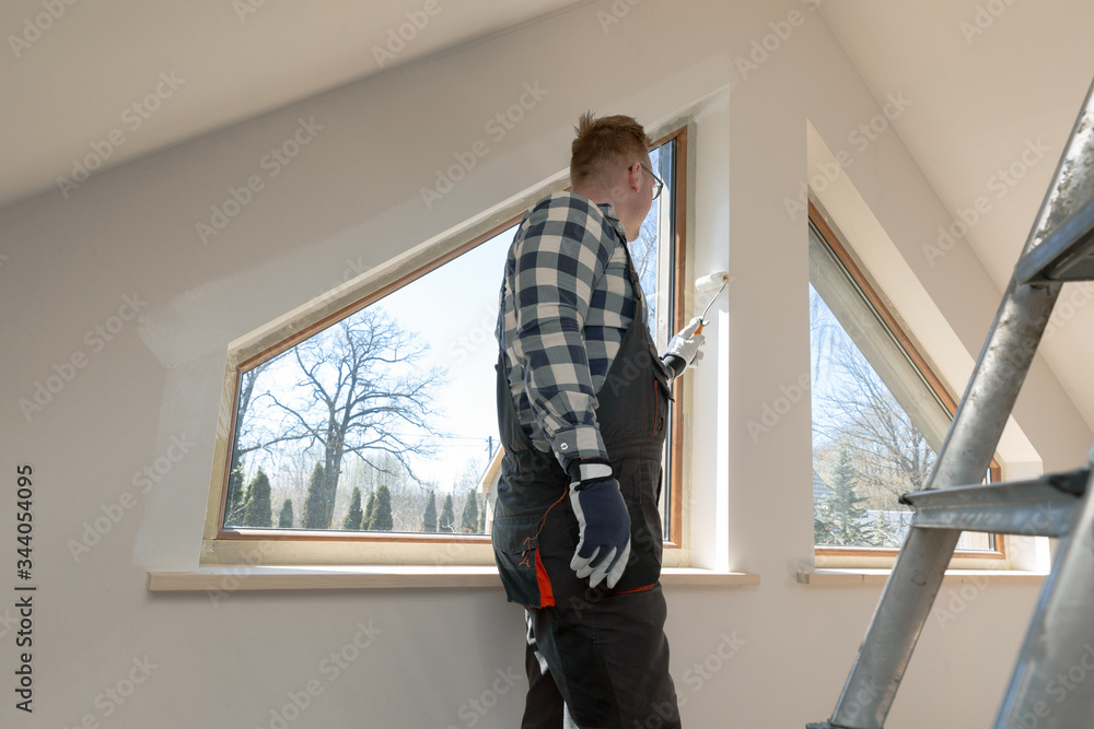 Home improvement concept, handyman painting a wall with a white paint near roof window in attic.