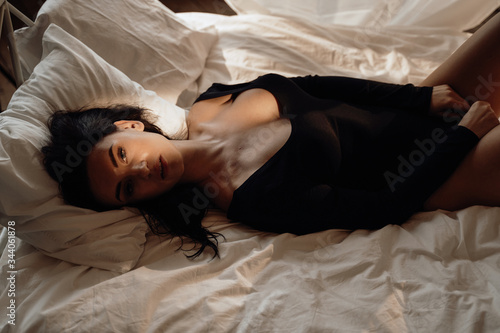 Graceful and cute woman with dark hair wearing black body costume laying on a bed among white sheets