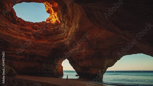Cinemagraph / seamless video loop of a young model woman in a dress standing on the beach in the famous Benagil Cave at Algarve, Portugal at sunrise in summer with the waves of the sea moving gently.