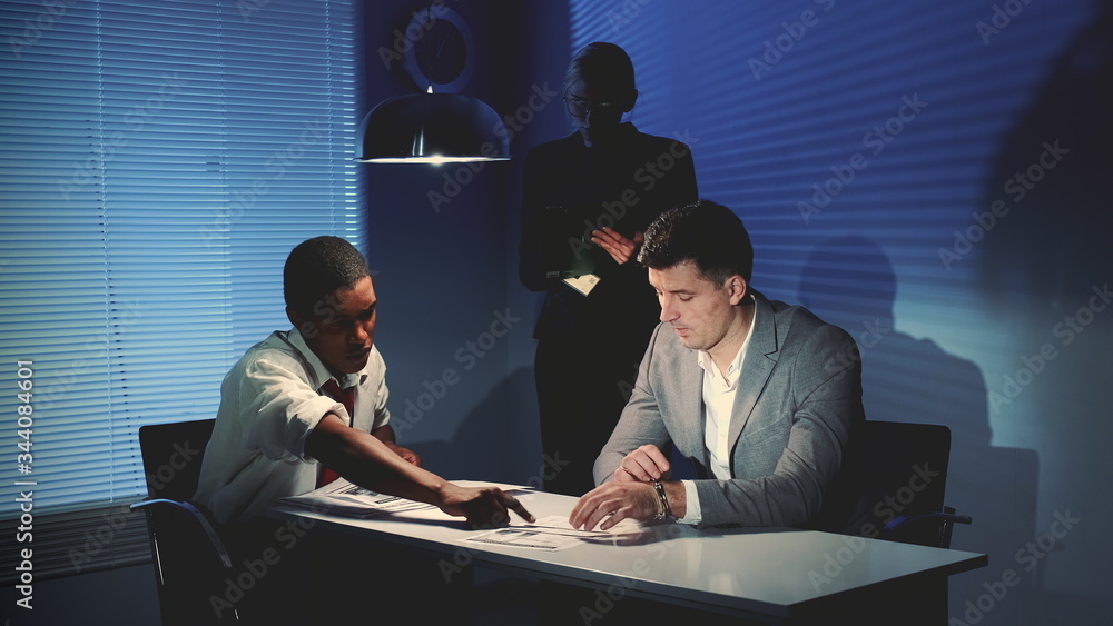 Medium shot of African detective questioning suspect caucasian man in interrogation room. Female assistant making notes of the interrogation. - obrazy, fototapety, plakaty 