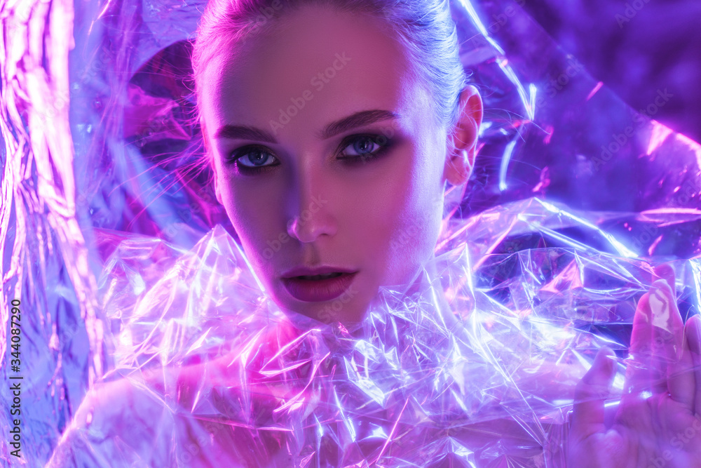 High Fashion model girl in colorful bright neon lights posing in studio through transparent film. Portrait of beautiful sexy woman in UV. Art design colorful make up. On colourful vivid background.