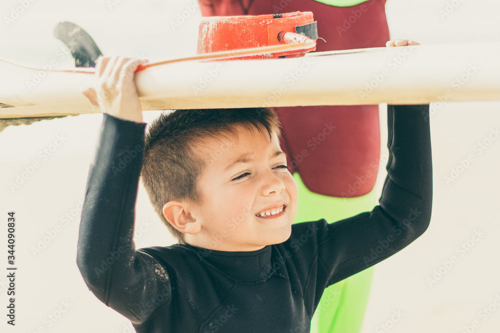 Cute little boy holding surfboard. Adorable smiling child in wetsuit holding surfboard and looking aside on sandy beach. Surfing concept Cute little boy holding surfboard. Adorable smiling child in wetsuit holding surfboard and looking aside on sandy beach. Surfing concept
