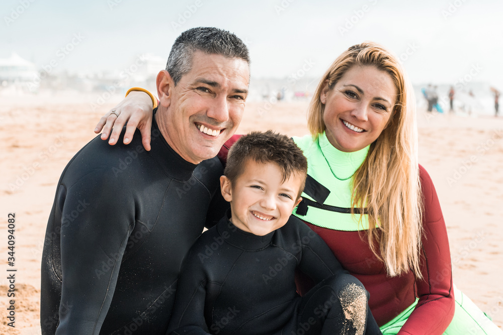 Happy family in wetsuits smiling at camera. Cheerful mother, father and son sitting together near surfboard on sandy beach. Surfing concept