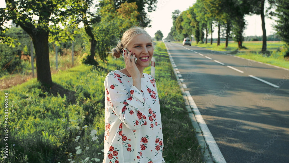 Young lady speaking on phone while hitchhiking. Side view of lovely young female talking on smartphone and waiting for car while standing on roadside during trip through countryside. Young lady speaking on phone while hitchhiking. Side view of lovely young female talking on smartphone and waiting for car while standing on roadside during trip through countryside.