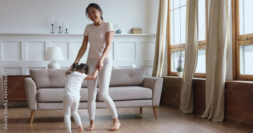 Full length overjoyed young asian woman holding hands of adorable little baby daughter, twisting having fun in modern living room. Happy vietnamese babysitter playing with cute small kid girl indoors.