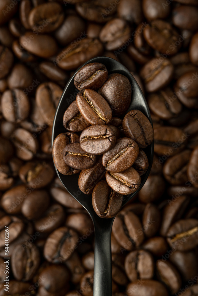 Coffee beans in black metal spoon in the foreground over a background of coffee beans