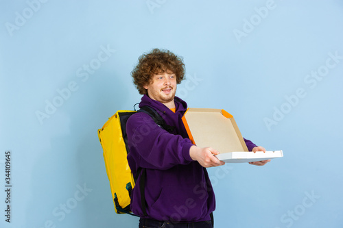 Proposing pizza to client, smiling. Emotions of caucasian deliveryman on blue background. Contacless delivery service during quarantine. Man delivers food during isolation. Safety. Hurrying up. photo