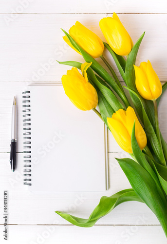 Beautiful yellow tulips and empty paper notebook, pen on white wooden background.
