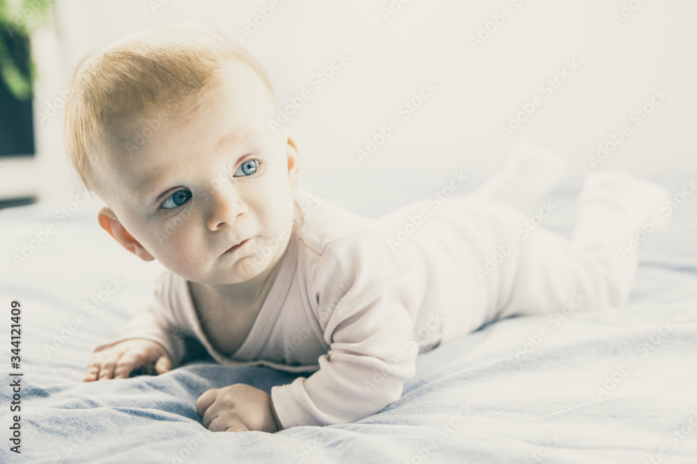 Serious pensive cute baby girl crawling in bedroom. Six month child lying on belly at home. Childhood or baby care concept
