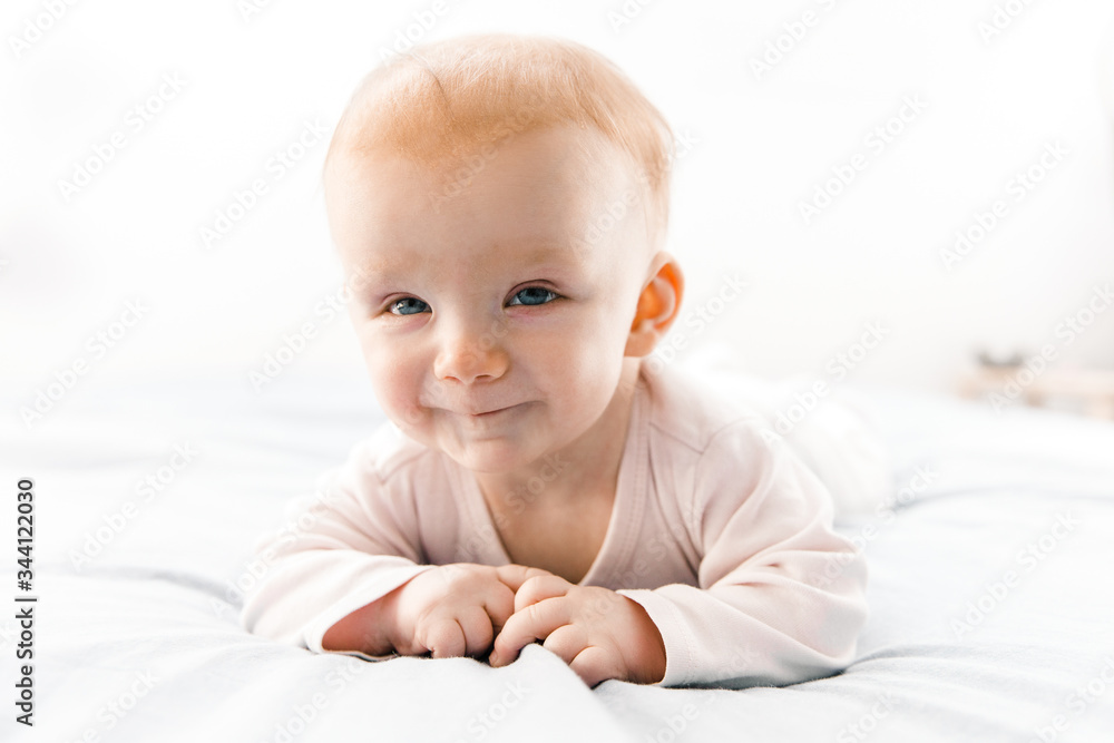 Joyful smiling red haired baby girl crawling on bed sheet and looking at camera. Six month child lying on belly in bedroom. Childhood or baby care concept
