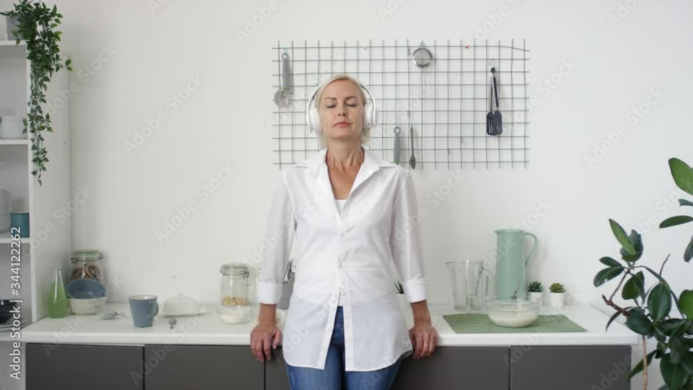 Beautiful middle aged woman standing with eyes closed beside kitchen counter, listening to music with wireless headphones and then looking at camera and smiling Beautiful middle aged woman standing with eyes closed beside kitchen counter, listening to music with wireless headphones and then looking at camera and smiling