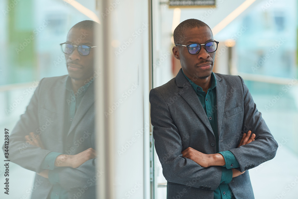 Portrait of African young businessman in suit and in eyeglasses standing with arms crossed and looking at camera at office Portrait of African young businessman in suit and in eyeglasses standing with arms crossed and looking at camera at office
