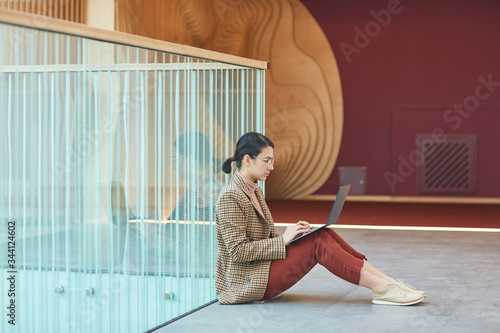 Young businesswoman sitting on the floor of office corridor and working online on laptop computer