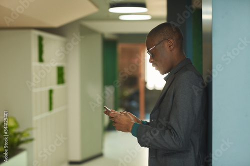 Young African businessman in suit standing at office corridor and typing a message on mobile phone