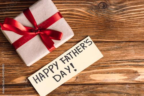 Top view of greeting card with lettering happy fathers day and pink gift box with red bow on wooden background