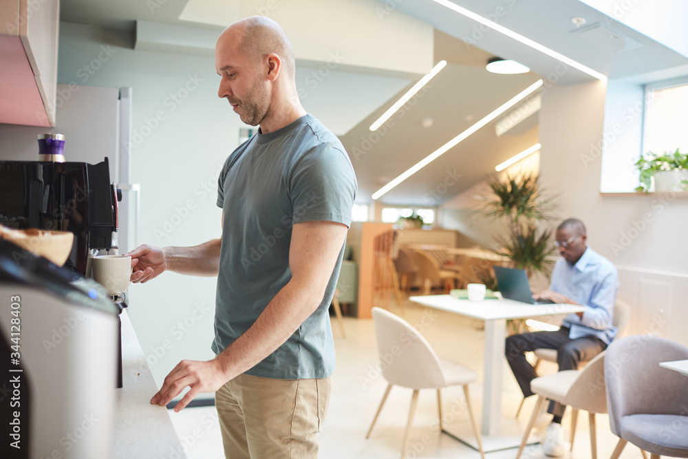 Mature man standing near the coffee machine and making coffee in the kitchen at office