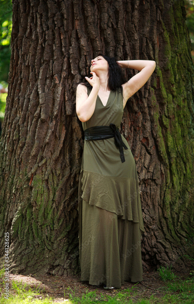 Young woman in green dress meditatively relaxes near large tree in forest park, concept of purity of nature and unity with environment Young woman in green dress meditatively relaxes near large tree in forest park, concept of purity of nature and unity with environment