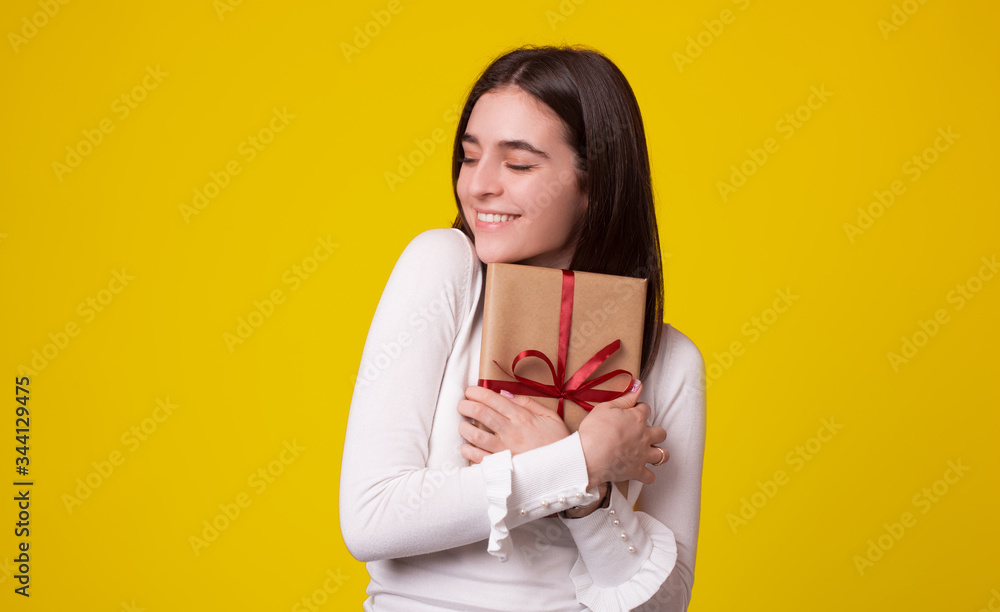 Fototapeta premium Cute girl is embracing a packed gift with eyes closed on yellow background.
