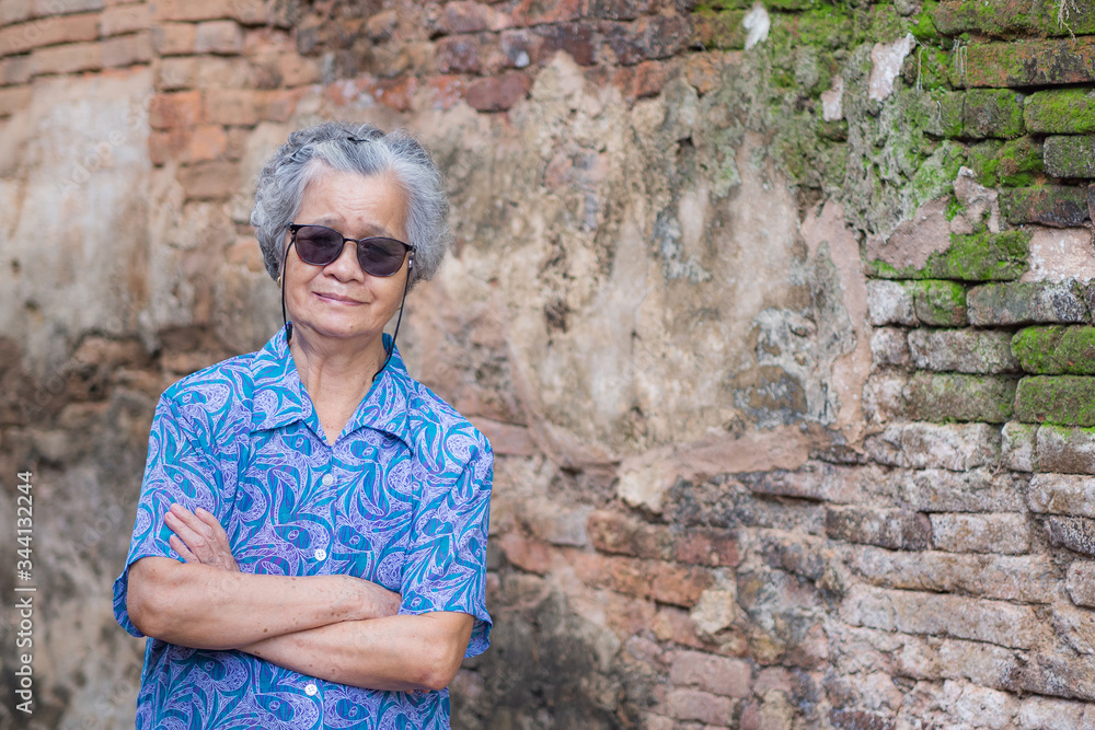 A portrait of an elderly woman wearing sunglasses, smiling and looking at the camera while standing arms crossed with old brick wall background. Space for text. Concept of old people and travel