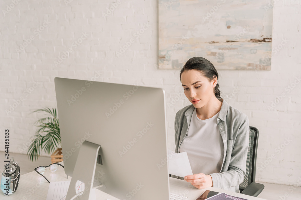 young, attentive freelancer looking at document while sitting near computer monitor young, attentive freelancer looking at document while sitting near computer monitor