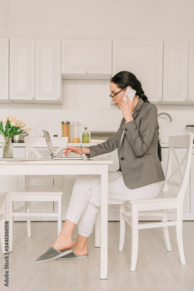 side view of businesswoman in blazer over pajamas working in kitchen, talking on smartphone and using laptop side view of businesswoman in blazer over pajamas working in kitchen, talking on smartphone and using laptop