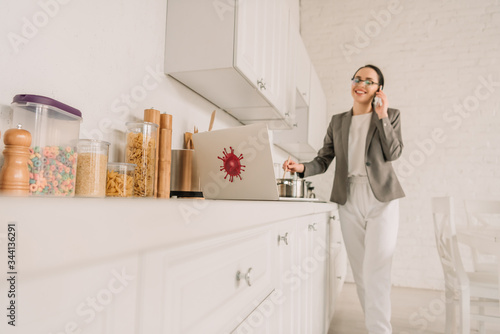 selective focus of businesswoman in a blazer over pajamas cooking and talking on smartphone in kitchen near laptop with coronavirus bacteria sticker