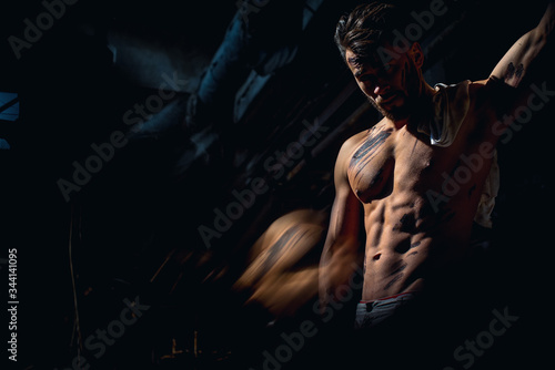 male athlete exhausted after hard work in factory, face in darkness, backlit muscles, bare torso, press and pumped muscles, t-shirt hanging on shoulders, look down.