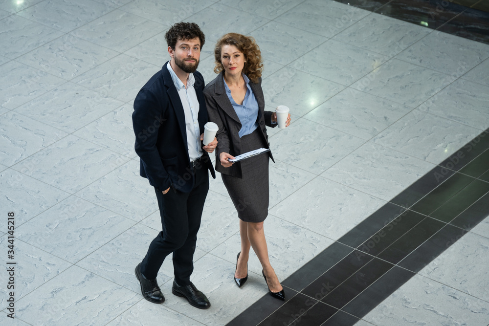 Business man and woman standing in the middle of the hall, drinking coffee and reading the document Business man and woman standing in the middle of the hall, drinking coffee and reading the document