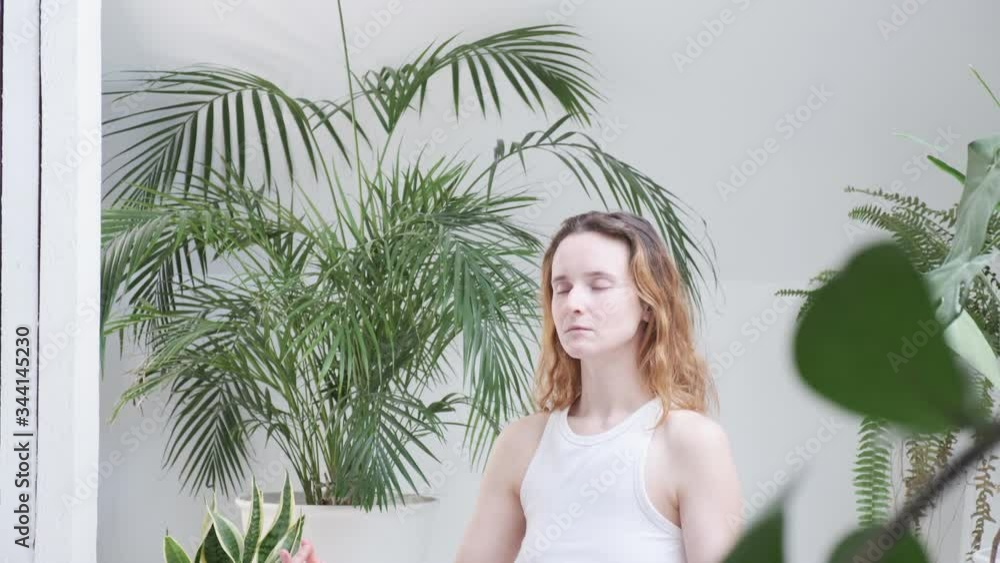 young red headed woman wearing sportswear closes eyes, starts meditating. girl sits in lotus pose, enjoys breath meditation in bright room with house plants at home during quarantine self isolation young red headed woman wearing sportswear closes eyes, starts meditating. girl sits in lotus pose, enjoys breath meditation in bright room with house plants at home during quarantine self isolation