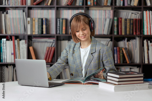 Cute young girl student working on a University project in the library on a laptop, with a book and headphones.