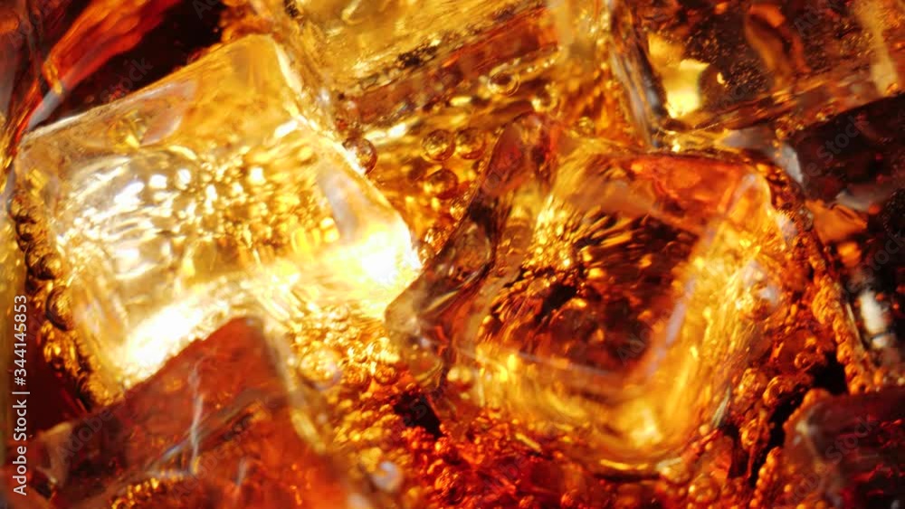 Ice cubes float and spin with bubbles in a glass of cola with brown highlights against black