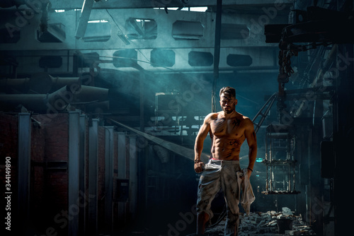 sporty bodybuilder confidently stands in metal production plant, shines side light, man with press and biceps, blue smoke on backlight illuminated by light, place for text.