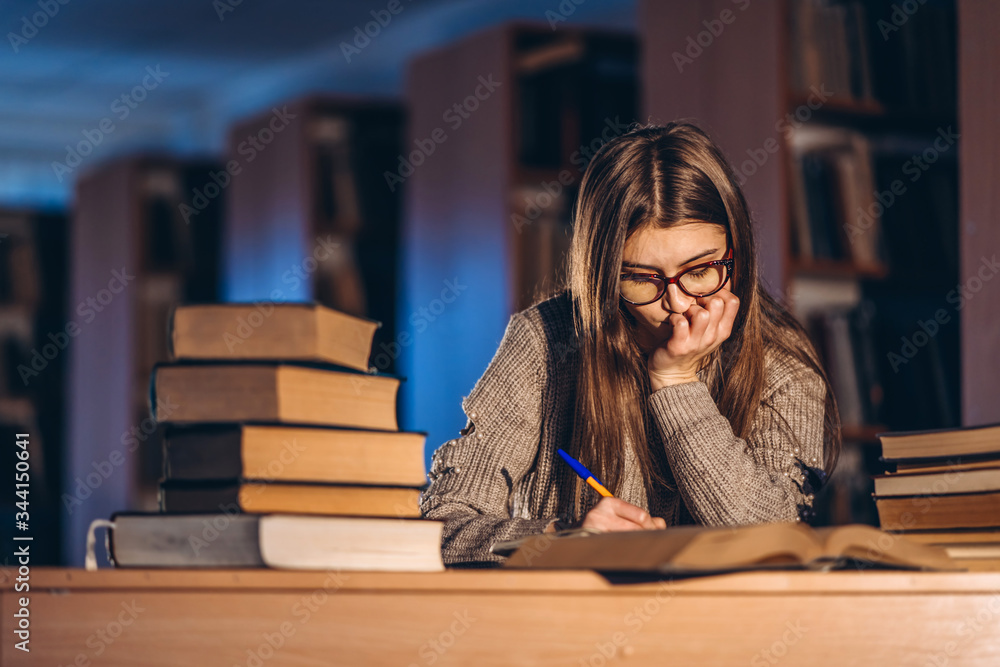 Young student in glasses preparing for the exam. Girl in the evening sits at a table in the library with a pile of books Young student in glasses preparing for the exam. Girl in the evening sits at a table in the library with a pile of books