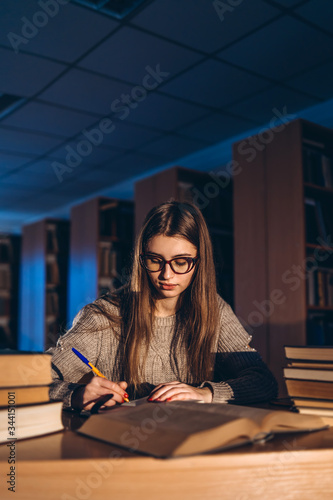 Young student in glasses preparing for the exam. Girl in the evening sits at a table in the library with a pile of books