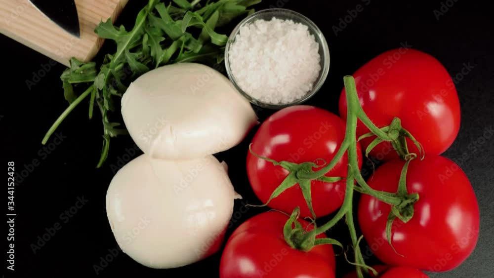 Modern caprese salad. Fresh juicy red tomatoes on a branch, mozzarella cheese, arugula leaves, olive oil and sea salt on a black background. Camera moves up to wooden board and knife. Top view. Close Modern caprese salad. Fresh juicy red tomatoes on a branch, mozzarella cheese, arugula leaves, olive oil and sea salt on a black background. Camera moves up to wooden board and knife. Top view. Close