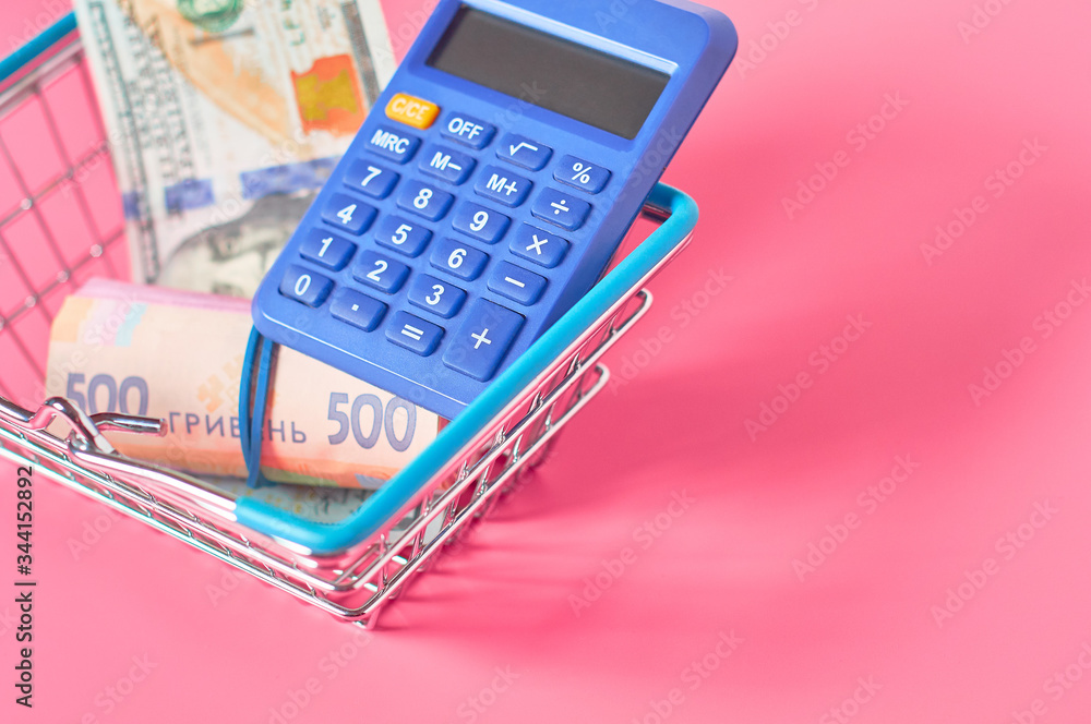 Different banknotes and blue calculator in metal market basket on pink background. Buy or sale money. Currency exchange concept Different banknotes and blue calculator in metal market basket on pink background. Buy or sale money. Currency exchange concept
