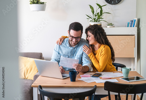Tablou pe pânză Positive young couple embracing and paying household bills or taxes on laptop on