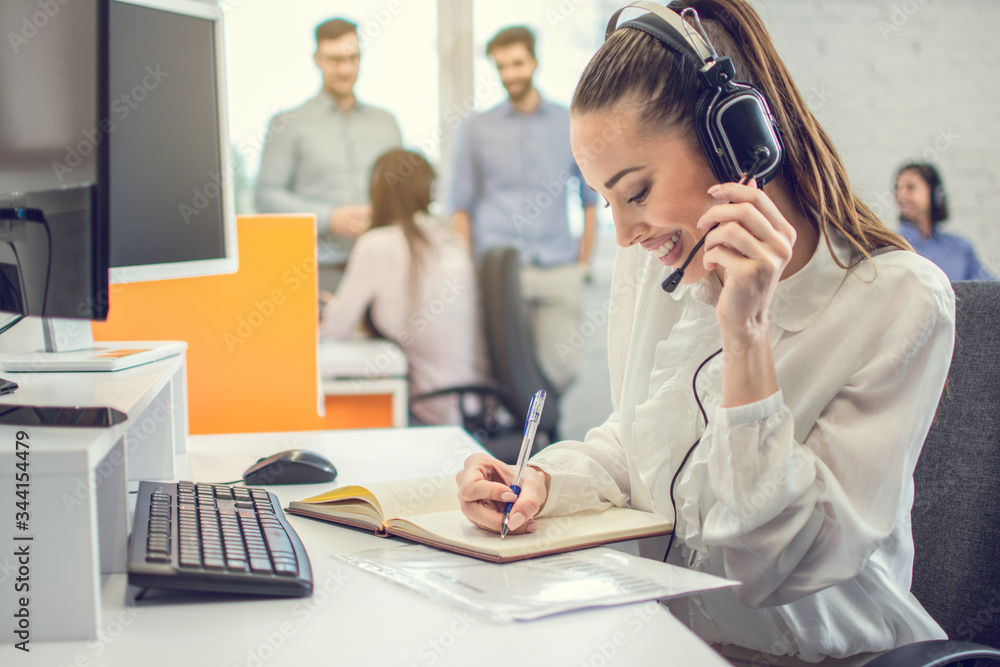 Attractive young business woman working in front of computer, writing notes to notebook at her workplace in office. Attractive young business woman working in front of computer, writing notes to notebook at her workplace in office.
