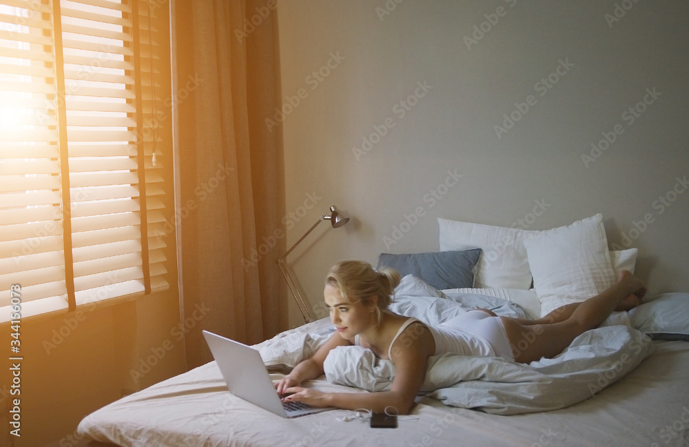 Sensual young woman in white tank top and panties lying on bed near laptop Sensual young woman in white tank top and panties lying on bed near laptop