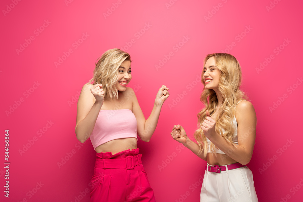 Cheerful blonde girls showing yeah gesture at each other on pink background Cheerful blonde girls showing yeah gesture at each other on pink background