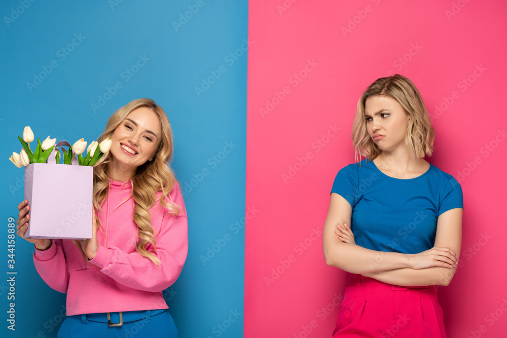 Smiling girl holding bouquet near envy blonde sister on blue and pink background - obrazy, fototapety, plakaty 