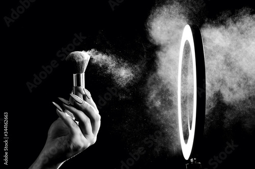 Make-up artist's hand with makeup brushes in front of a ring lamp against a dark background. Black and white photo