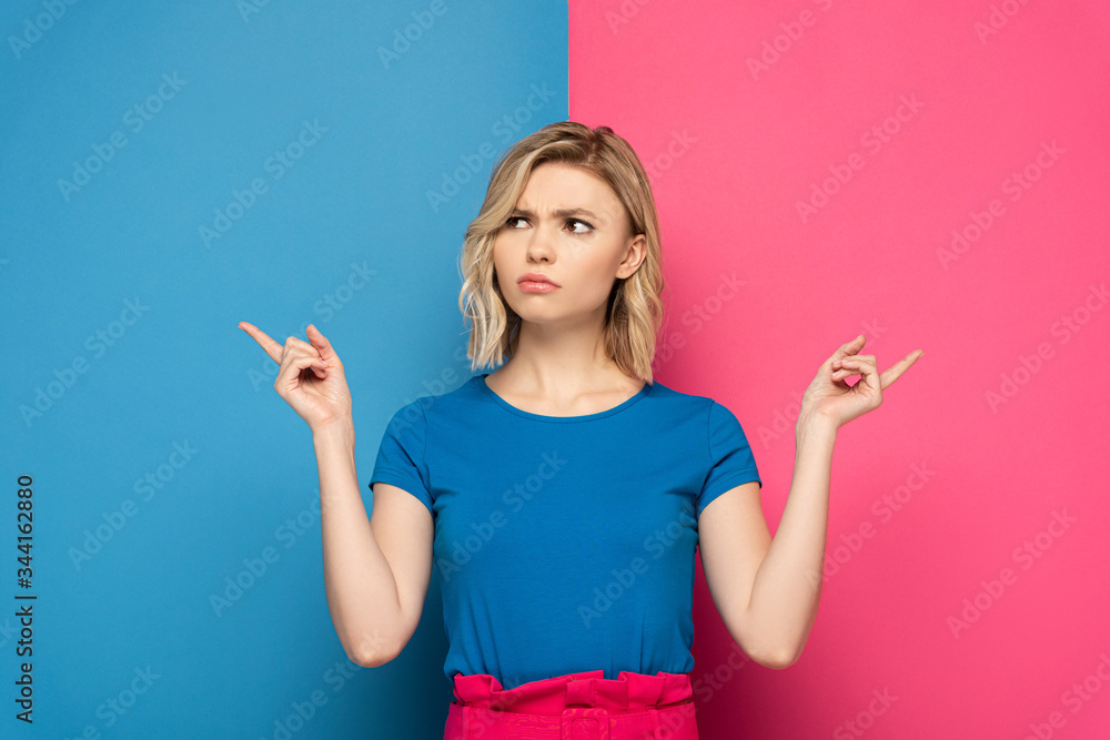 Pensive blonde woman pointing with fingers on pink and blue background Pensive blonde woman pointing with fingers on pink and blue background