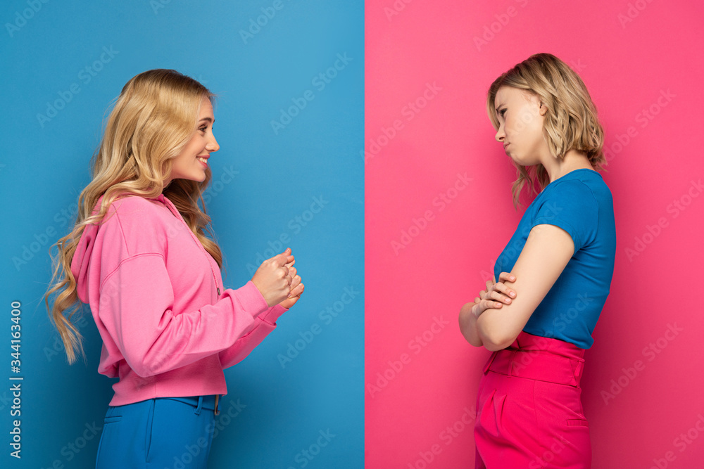 Side view of sad and happy blonde sisters looking at each other on pink and blue background Side view of sad and happy blonde sisters looking at each other on pink and blue background